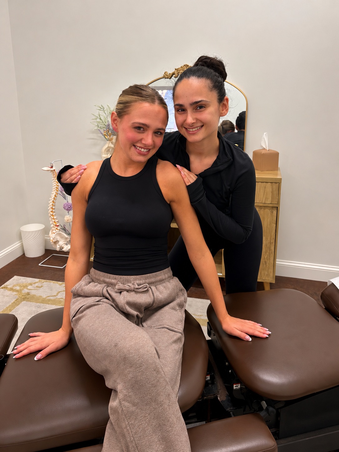 Two women smiling, one sitting on a massage table, the other standing behind her, in a room with a mirror and dresser.