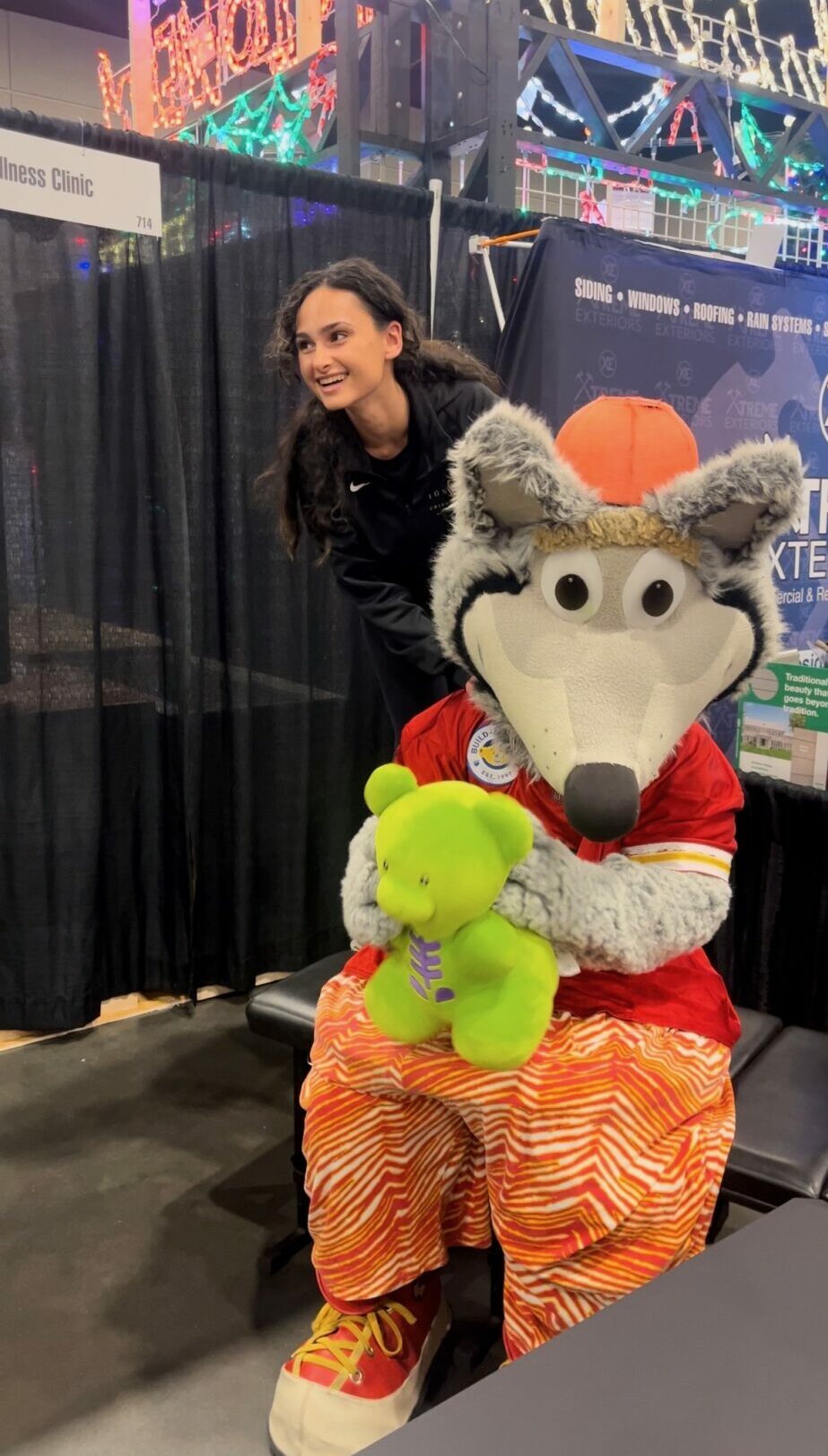 Person smiling with a costumed character holding a green plush toy at an indoor event, colorful lights in background.