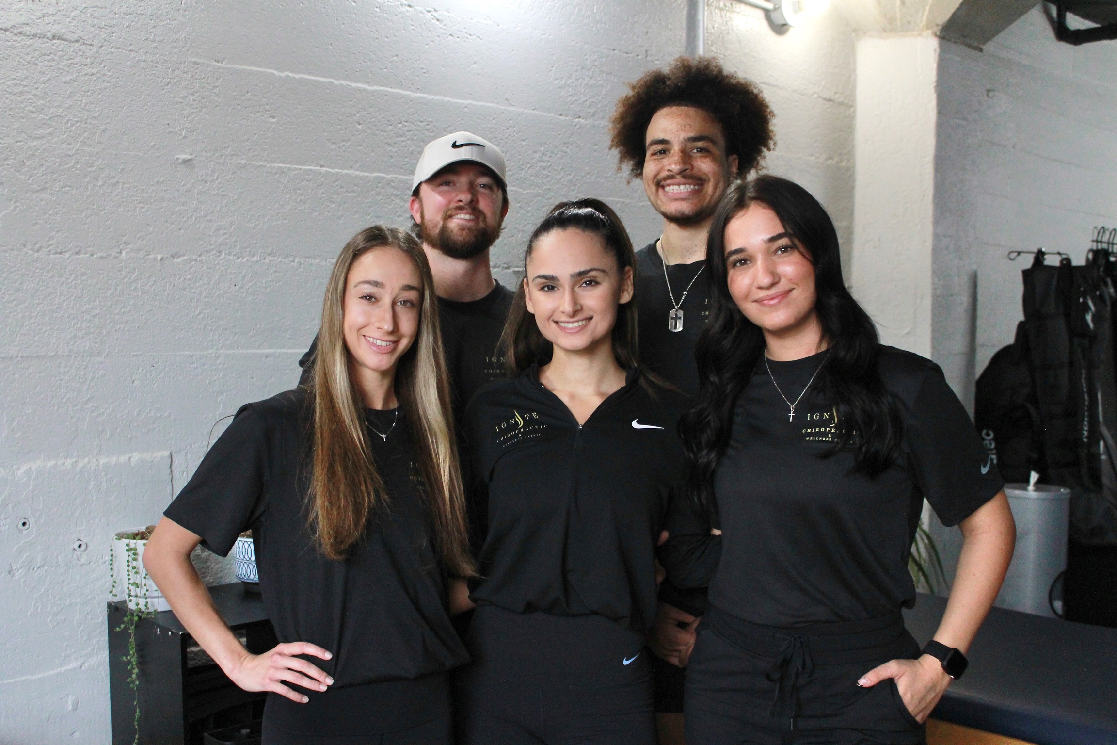 Group of five young people smiling, standing indoors against a gray wall, wearing black and dark clothing.