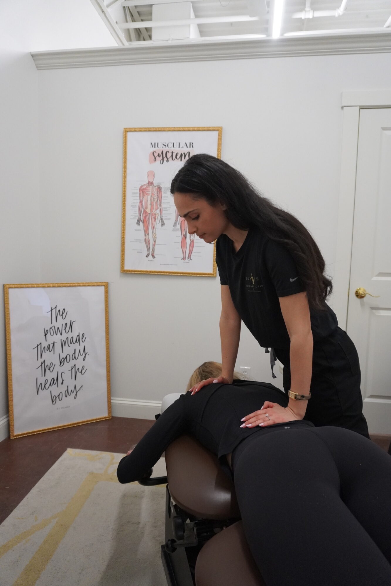 A woman provides a massage or therapy to a person lying face down on a massage table in a room with framed artwork on the wall.