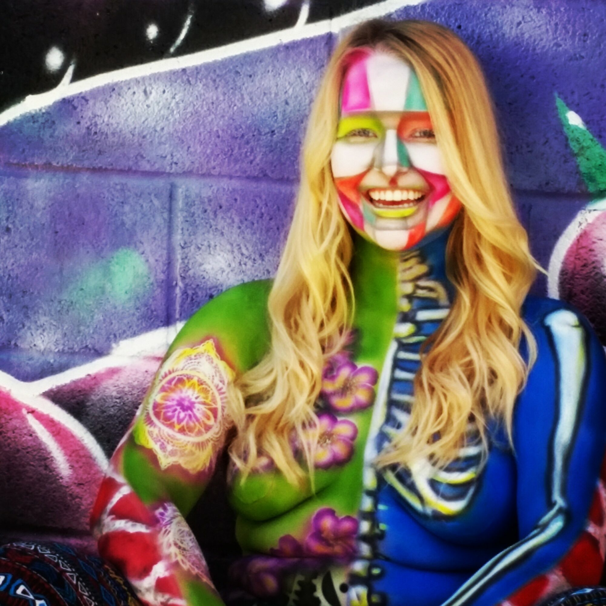 Woman with painted face and long blonde hair standing in front of a colorful graffiti wall.