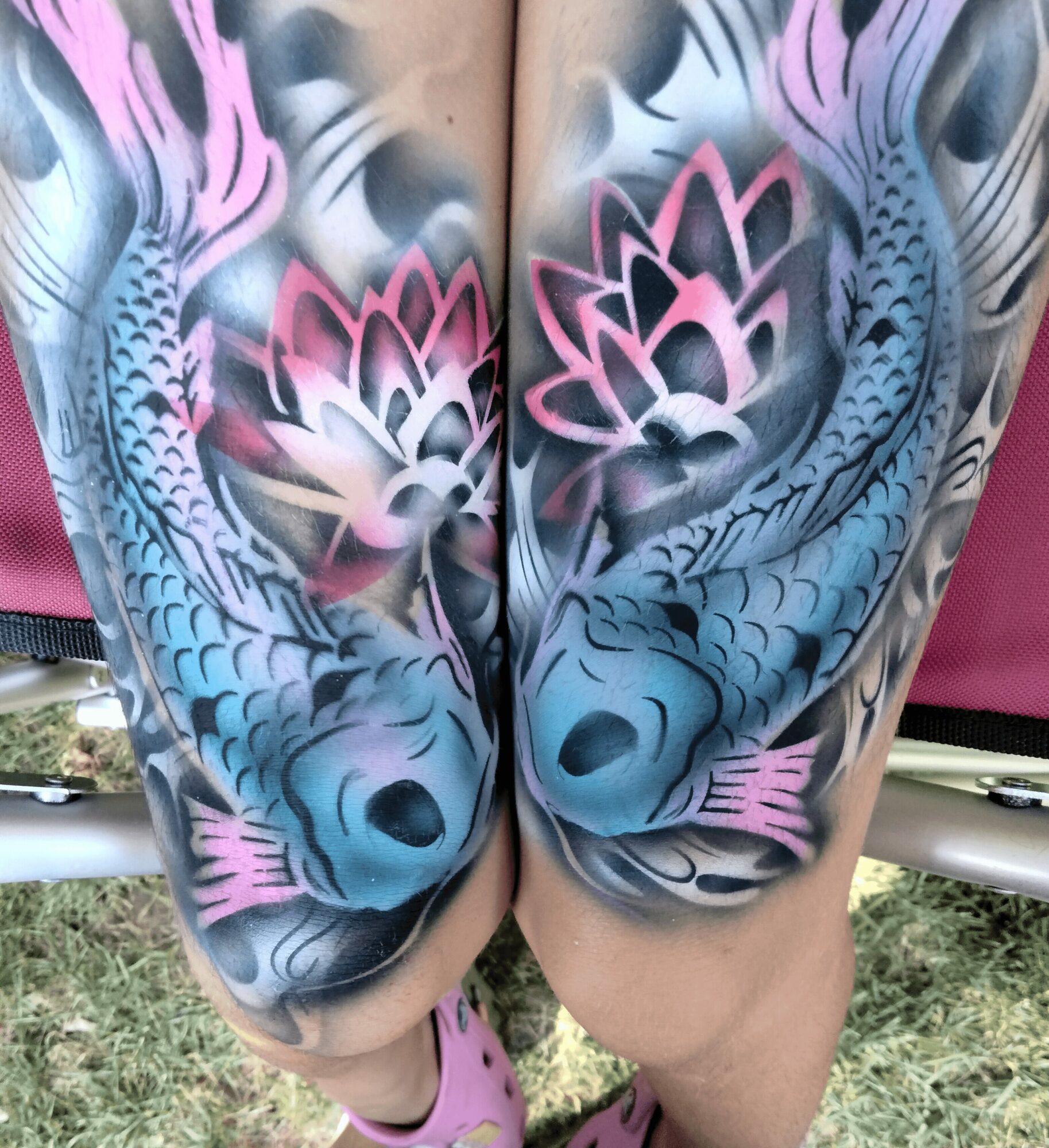 Tattoo of two colorful dragons with pink, blue, and black scales on person's thighs, outdoors on grass.