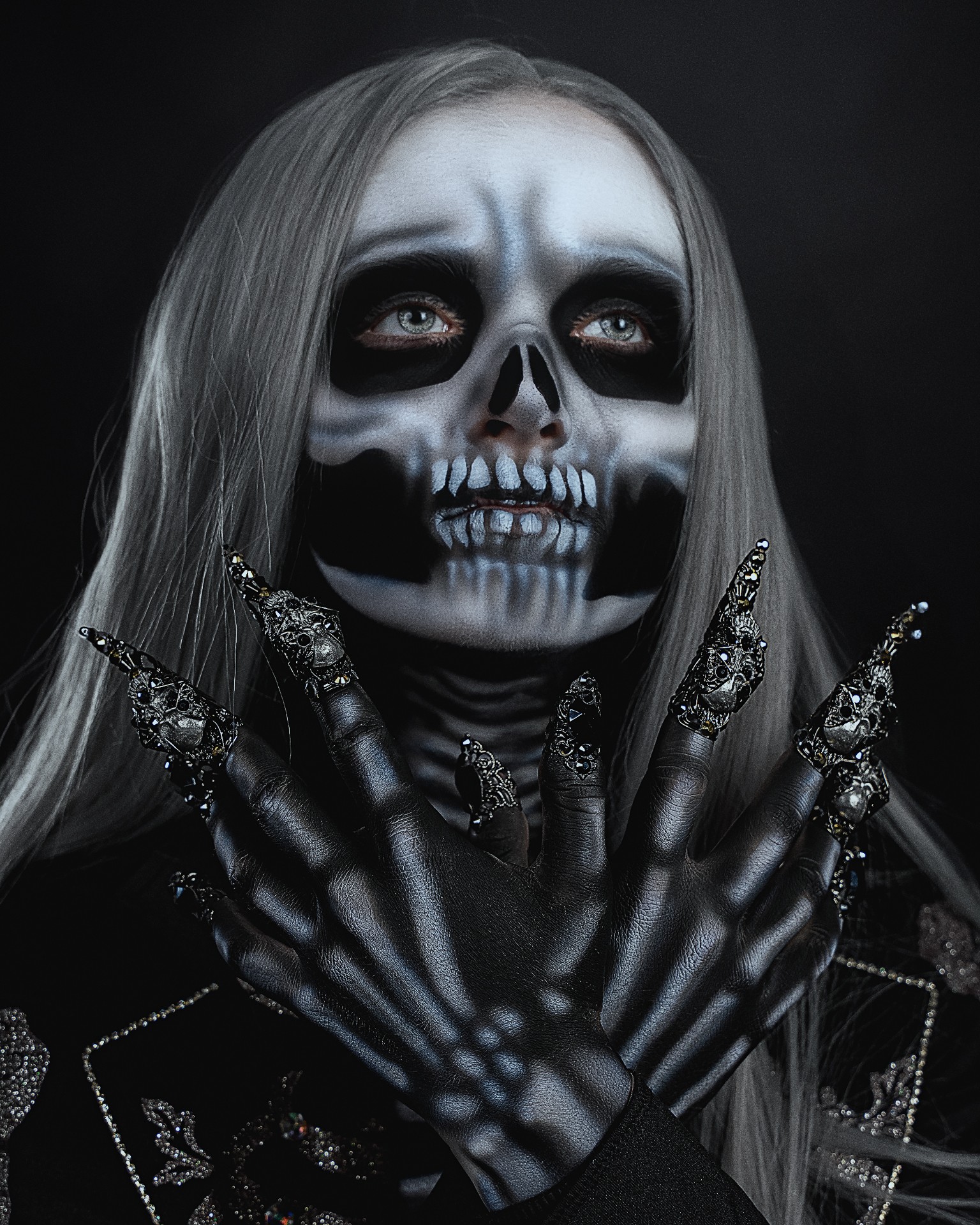 Person with skull makeup, long hair, dark clothing, and decorated black gloves, holding hands near face, dark background.