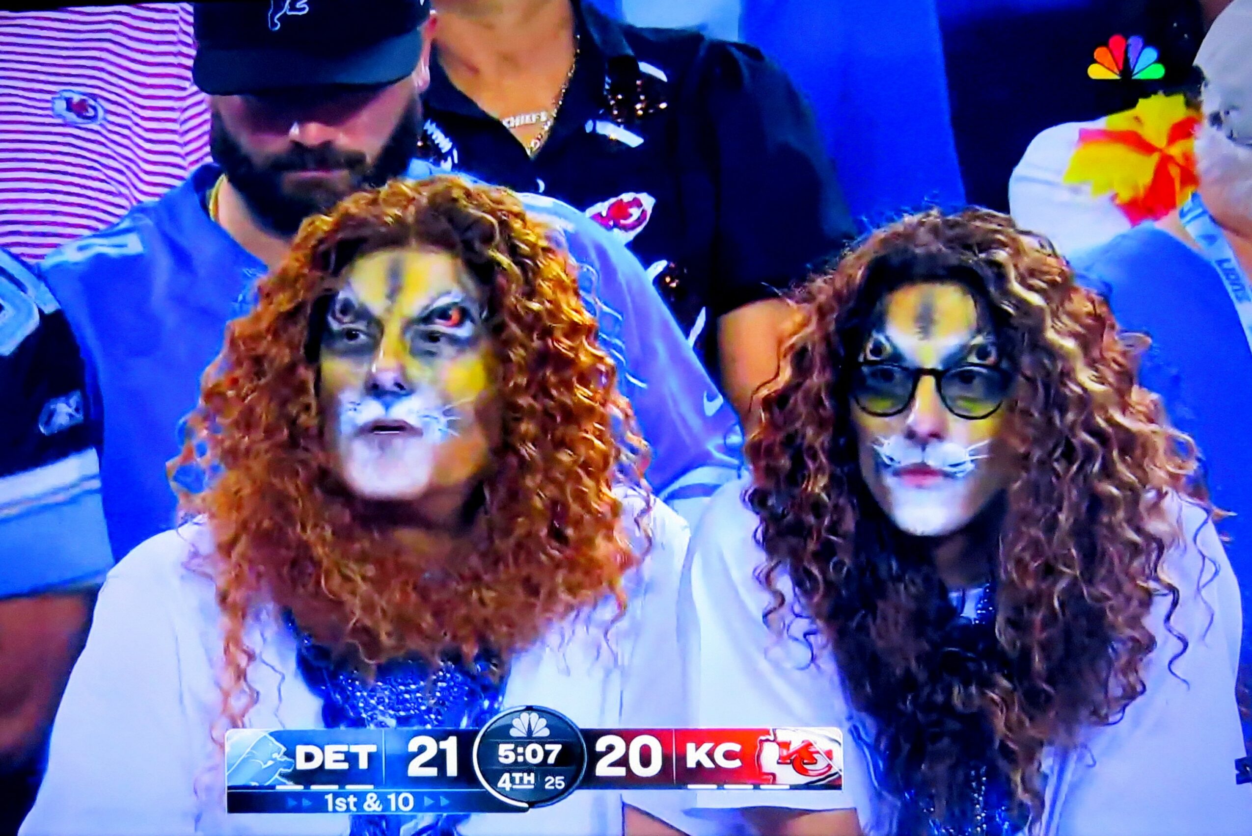 Two women with face paint resembling cats, curly hair, and wearing white shirts, sitting in a sports stadium.