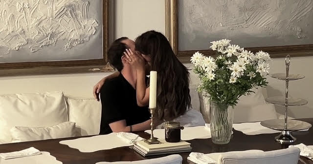 Two people kissing at a table with a large vase of white flowers, candles, and books, in a room with artwork.