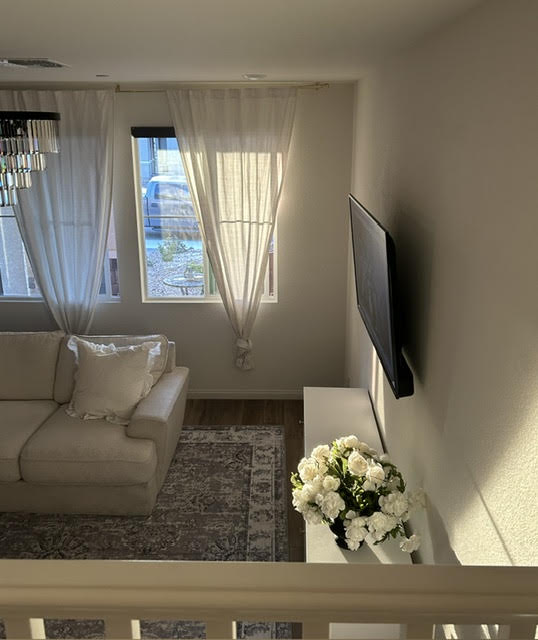 Living room with a sofa, window with curtains, wall-mounted TV, and a flower arrangement on a white table.