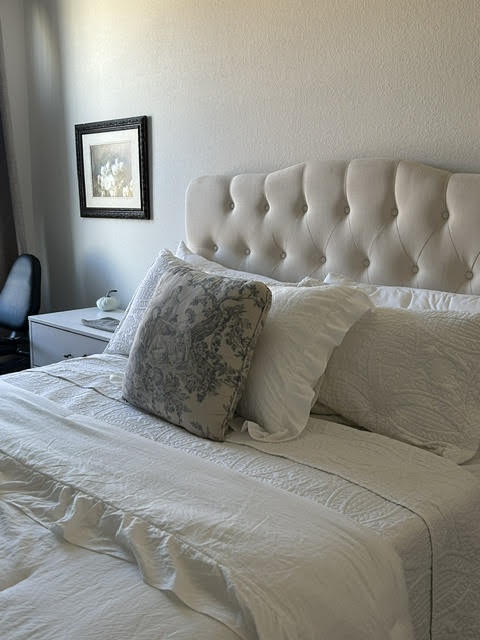 Bed with white bedding, decorative pillow, tufted headboard, nightstand, framed picture, and a chair in the corner.