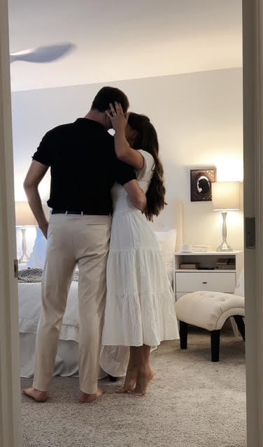 A couple stands close, touching foreheads and holding hands in a cozy room with a lamp and furniture.