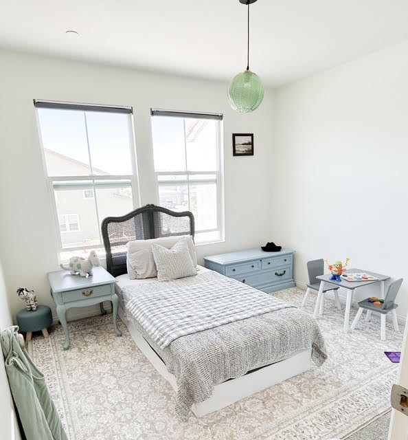 Bedroom with a bed, two windows, a nightstand, a dresser, and a small table with chairs, decorated in light colors.