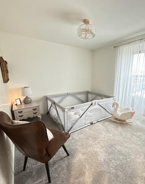 Room with a white crib, brown chair, nightstand with lamp, and large window with sheer curtains.