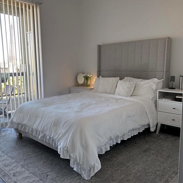 Bedroom with a bed, gray headboard, white bedding, nightstands, and a window with vertical blinds.