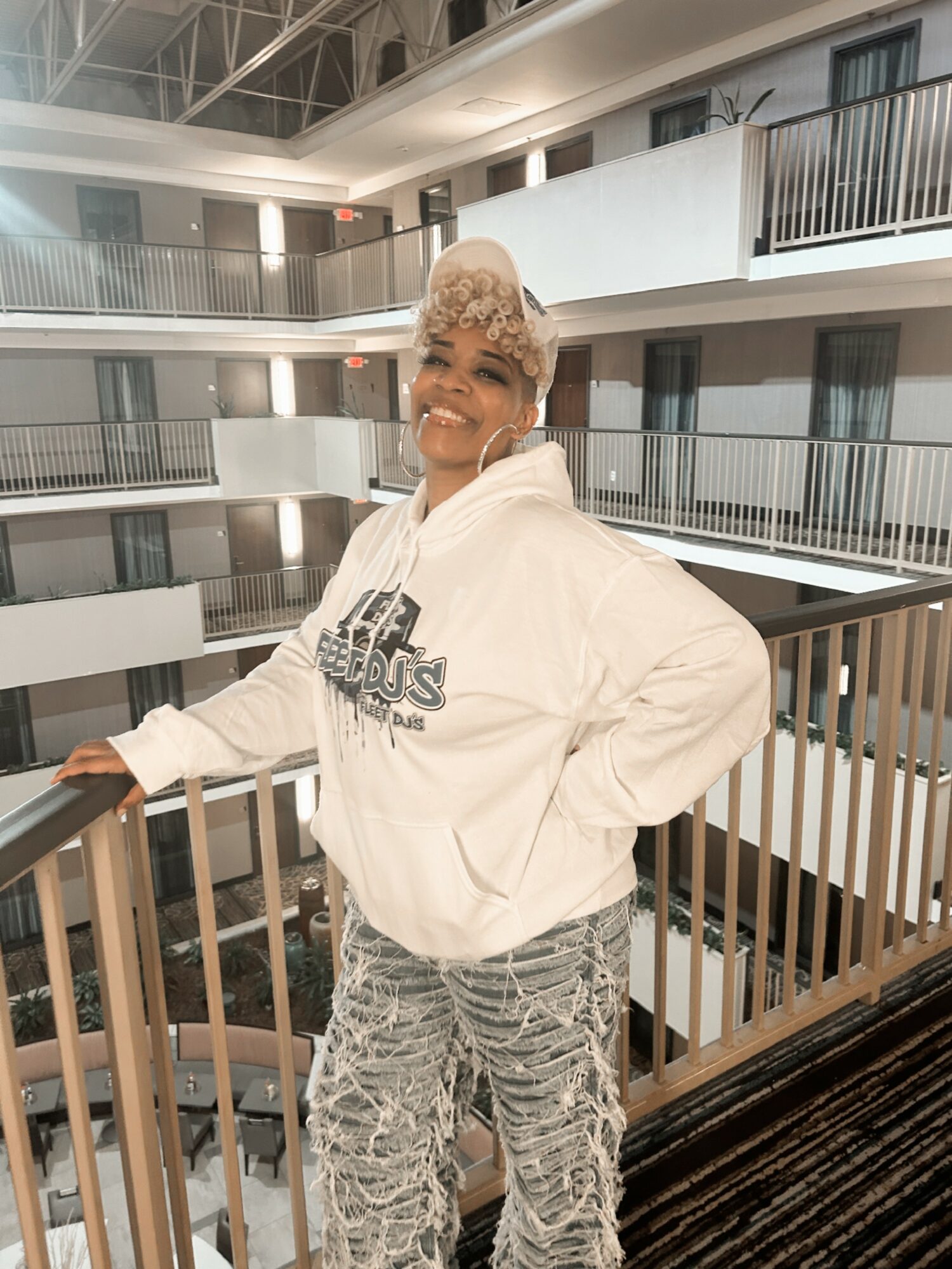 Smiling woman in a white hoodie and patterned pants, standing on a balcony inside a multi-story building.