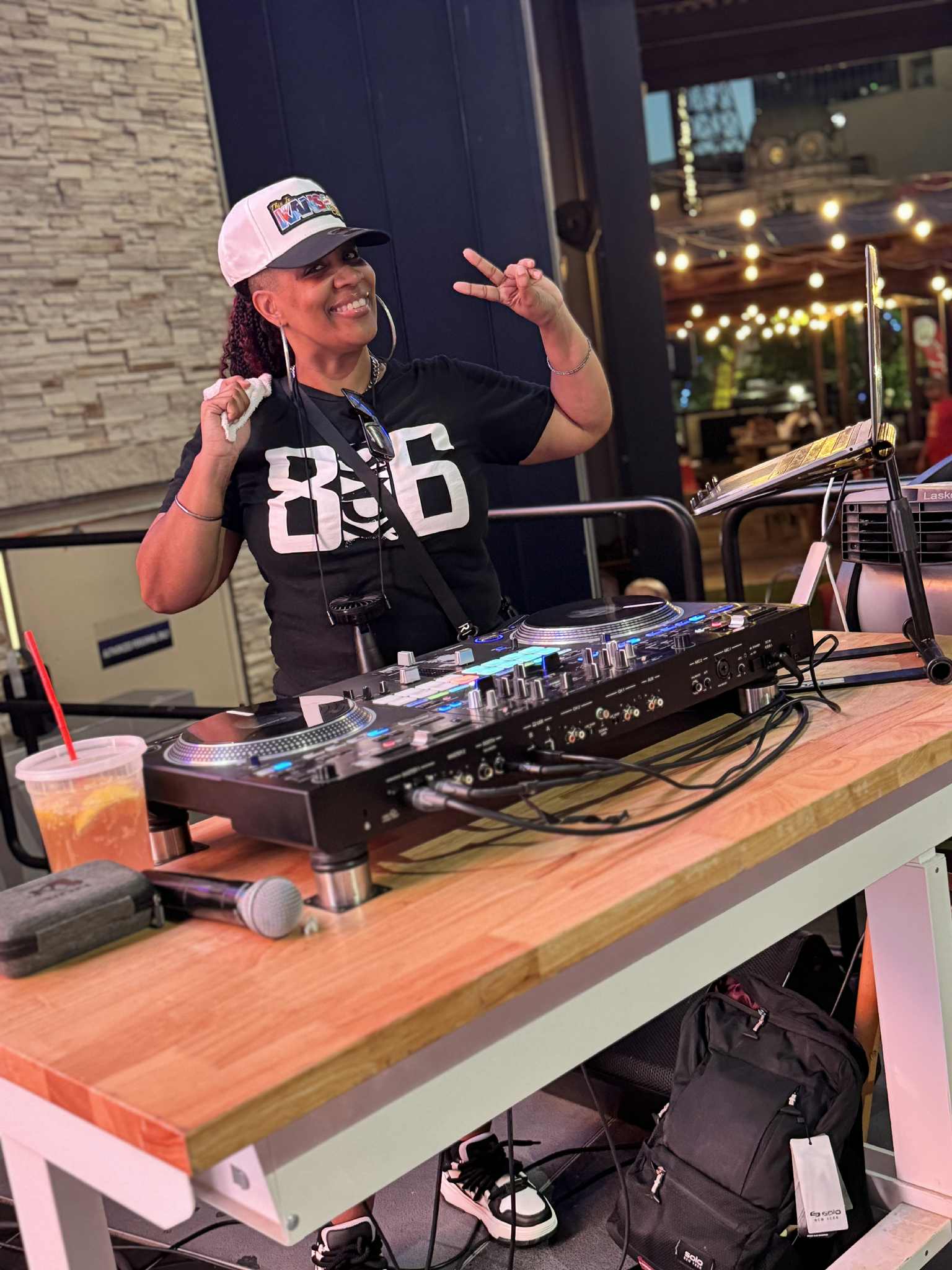 Person DJing with headphones, smiling, making a peace sign, standing behind a DJ mixer at a venue with string lights.