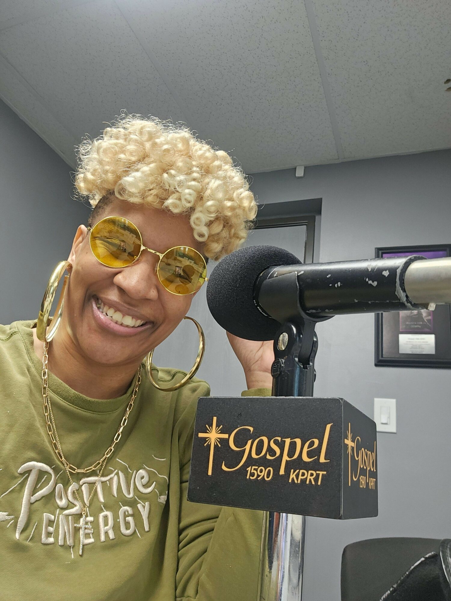 Person with curly blonde hair and yellow sunglasses smiling at a microphone during a press event.