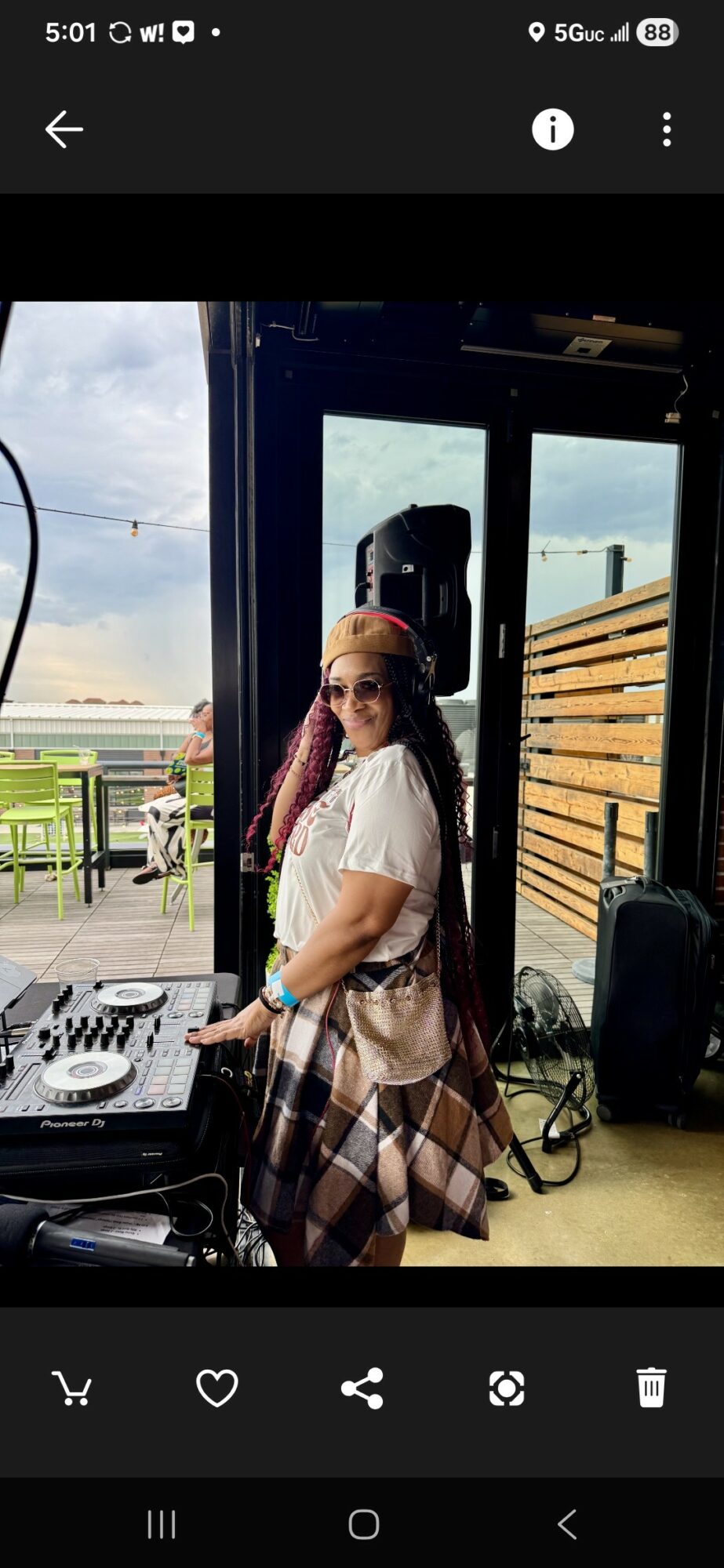 Woman with long hair wearing sunglasses, standing indoors near DJ equipment, with a window showing an outdoor scene.