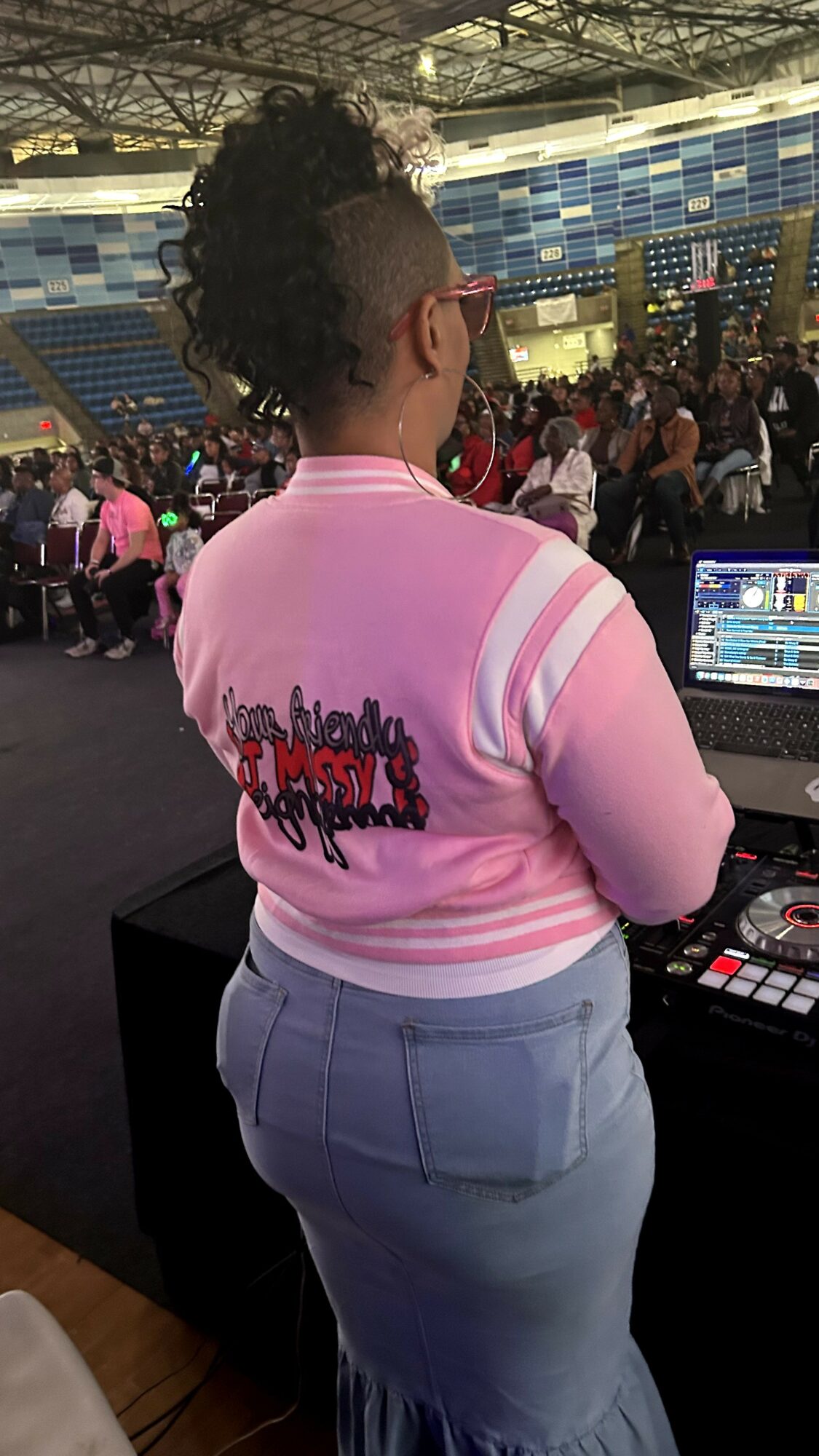 Person with curly hair and glasses wearing a pink and white striped shirt, standing at DJ equipment in a large indoor venue.