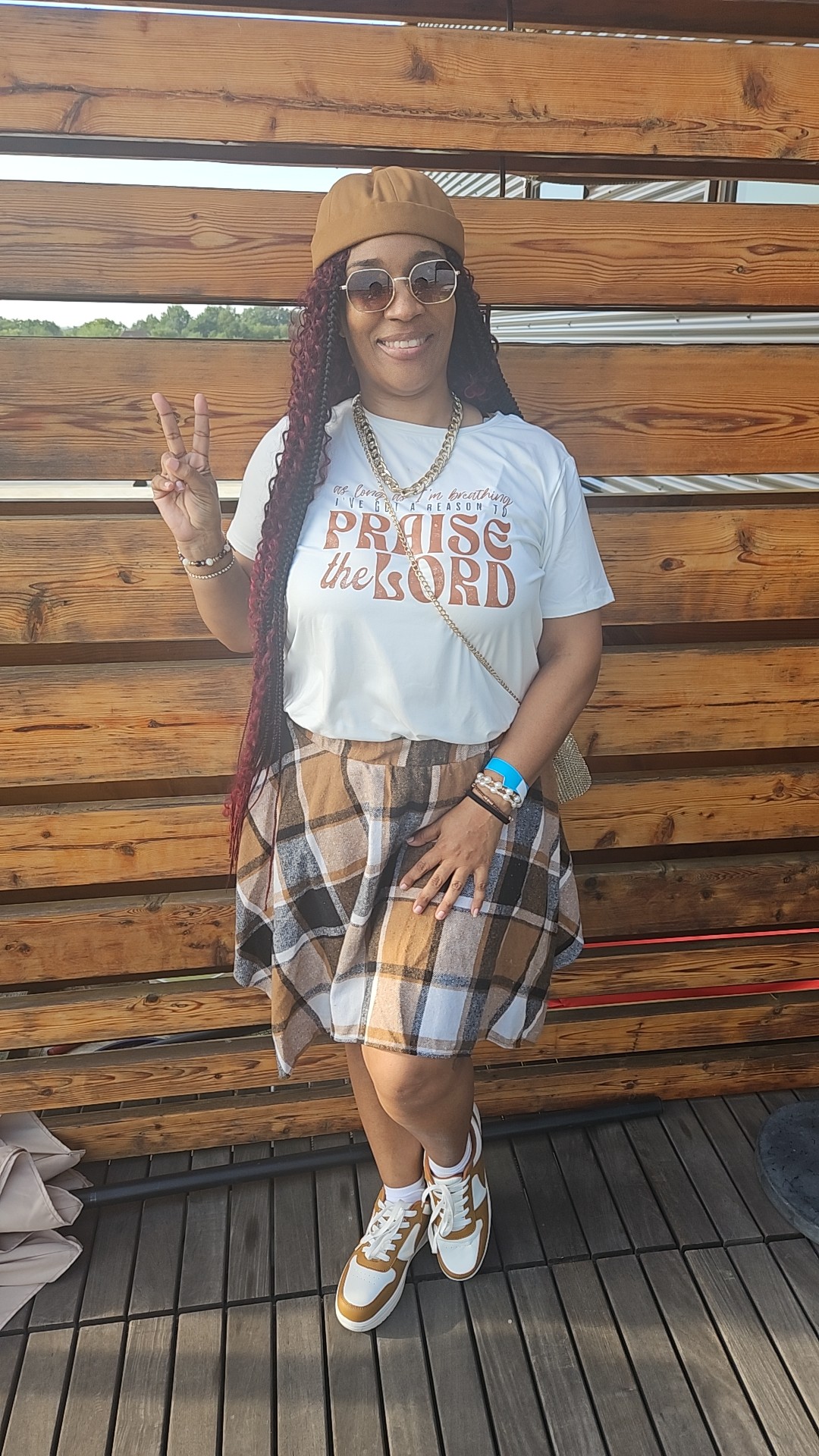 Woman with long braids, sunglasses, and a beanie, smiling and making a peace sign, standing outdoors against a wooden fence.