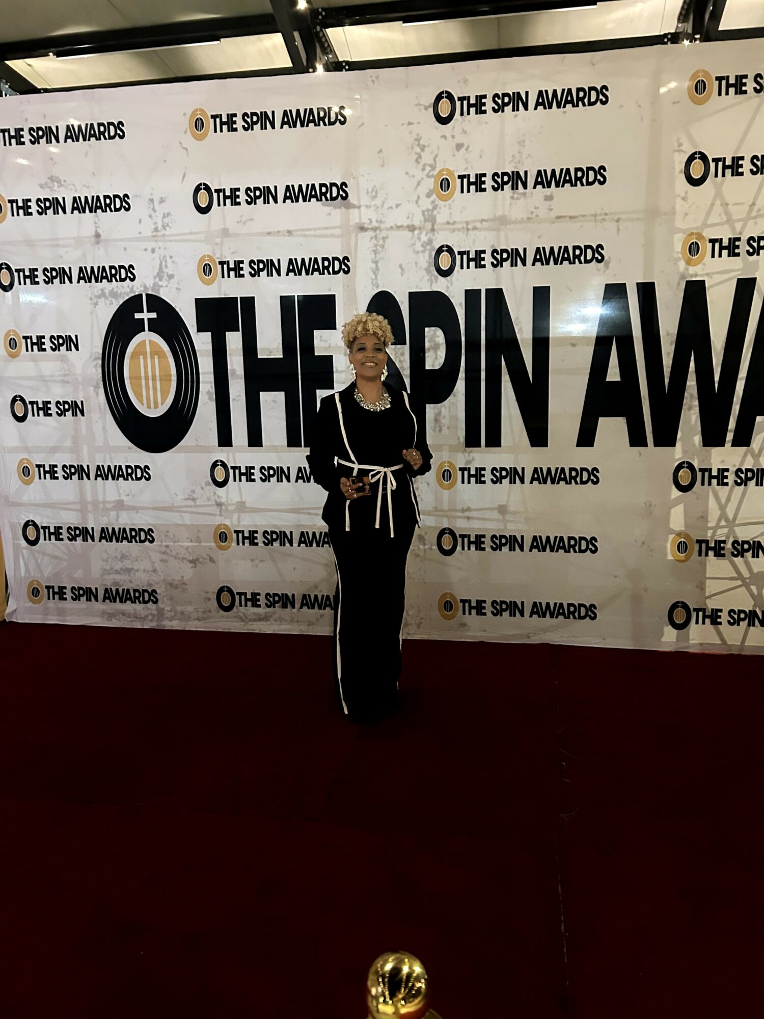 Person standing in front of a backdrop with 'The Spin Awards' text, wearing a black outfit with white accents, holding a trophy.
