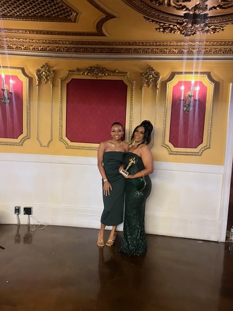 Two women in elegant dresses pose in a decorated room with ornate gold and red wall panels, and a chandelier ceiling.