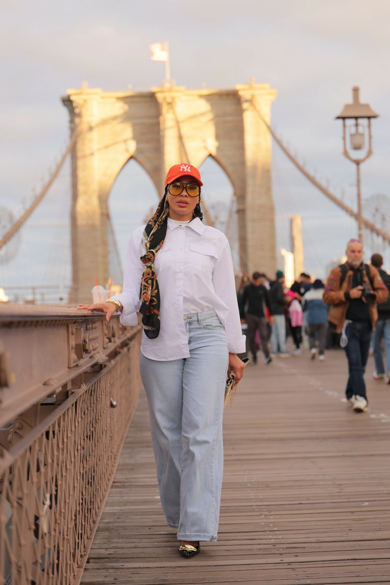 Woman in white shirt and jeans standing on a bridge with Brooklyn Bridge in background, people walking nearby.