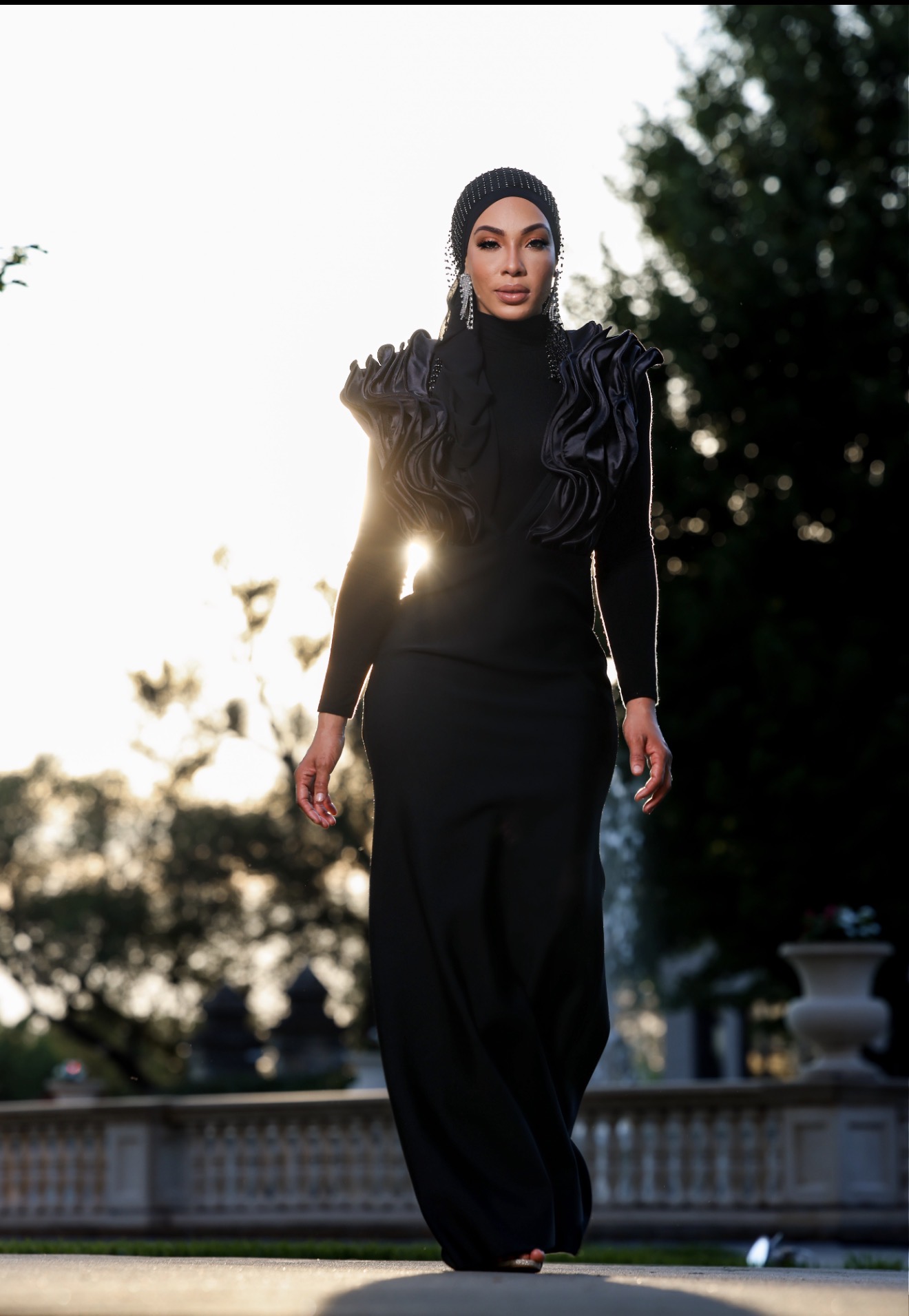 Woman in black dress and hijab walking outdoors with trees and a fence in background.
