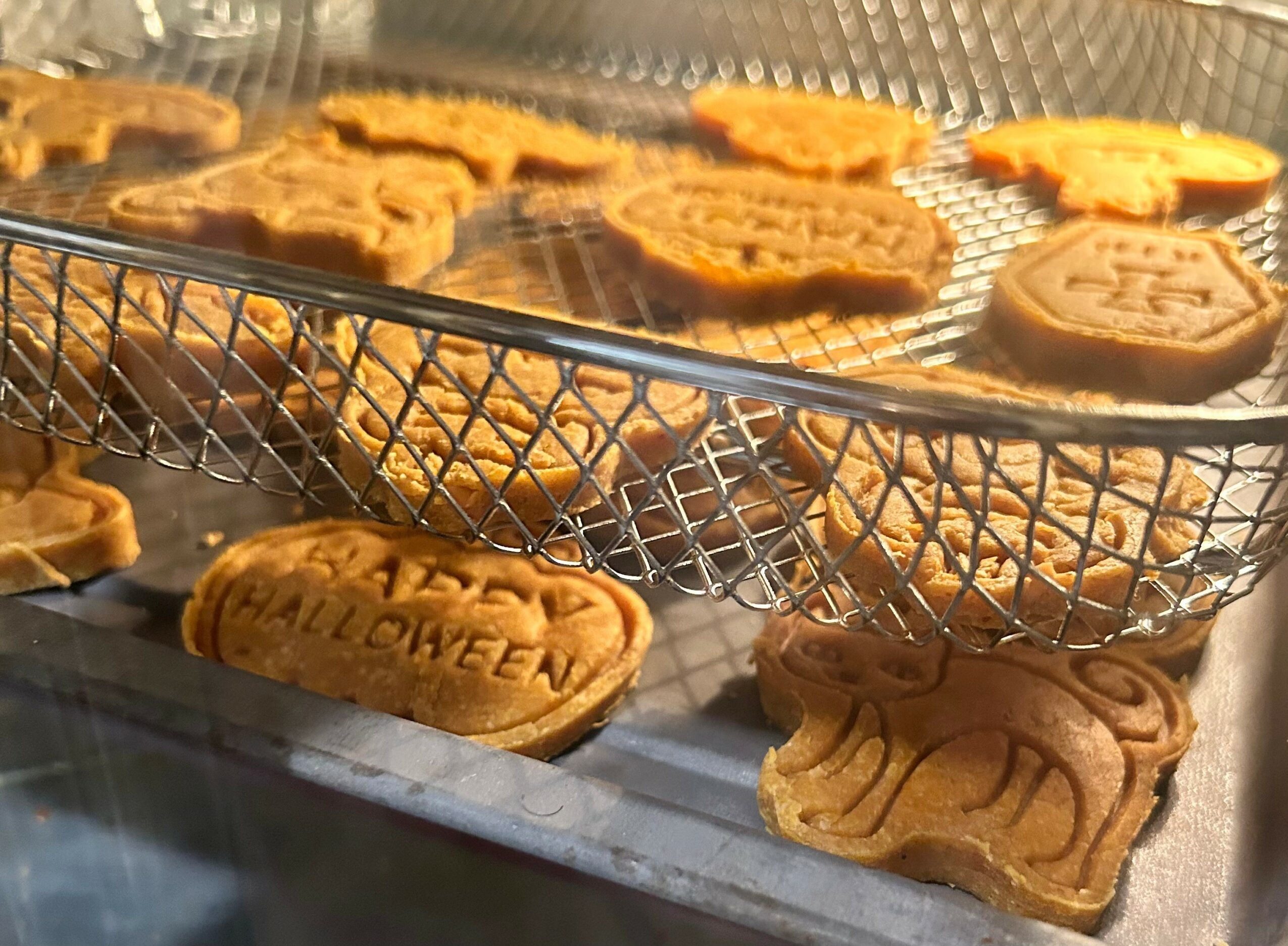 Cookies on a wire rack and tray, some with Halloween and other designs, in a baking setting.