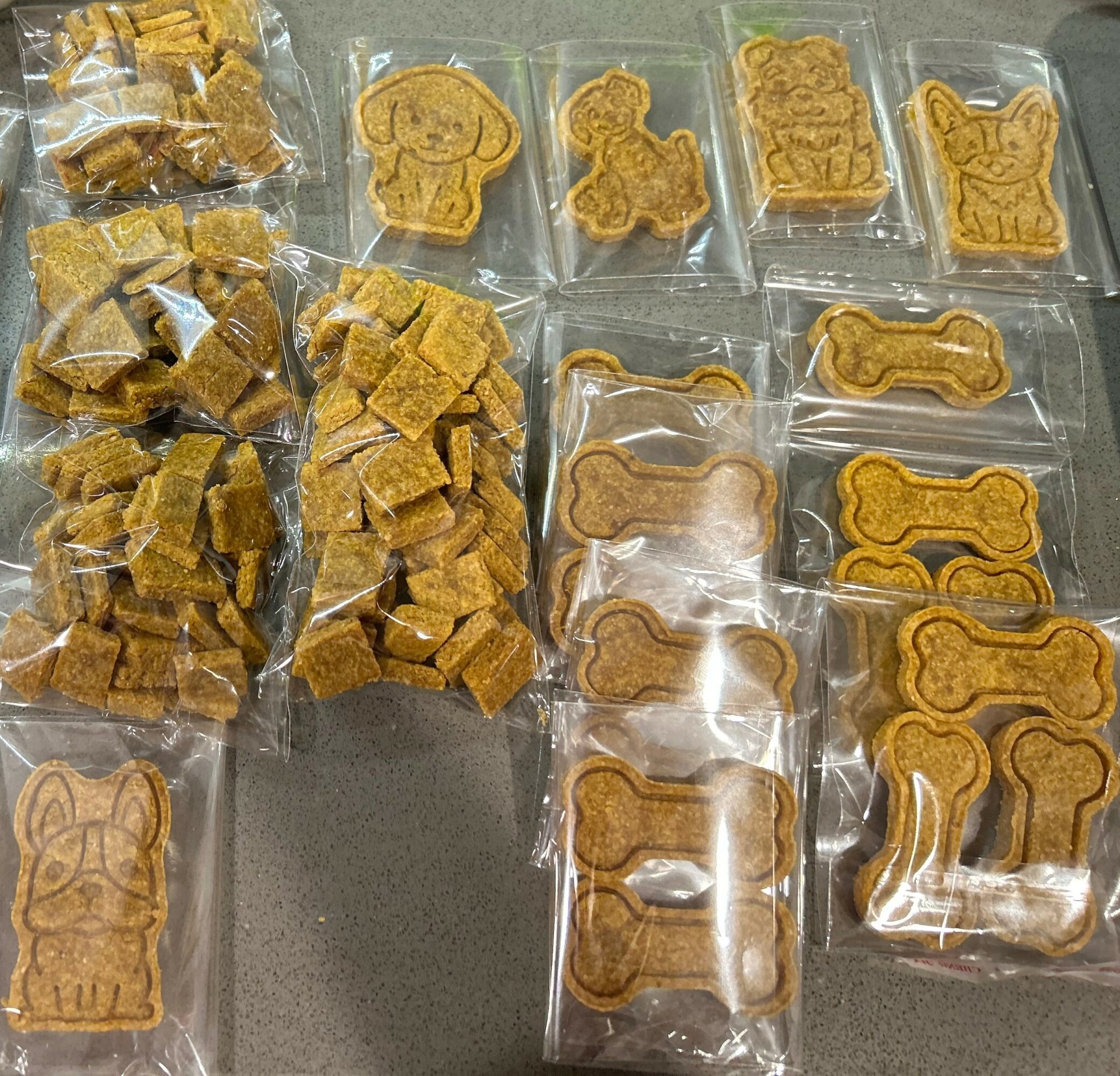 Assorted dog-shaped cookies and small snack pieces in clear plastic bags on a gray surface.