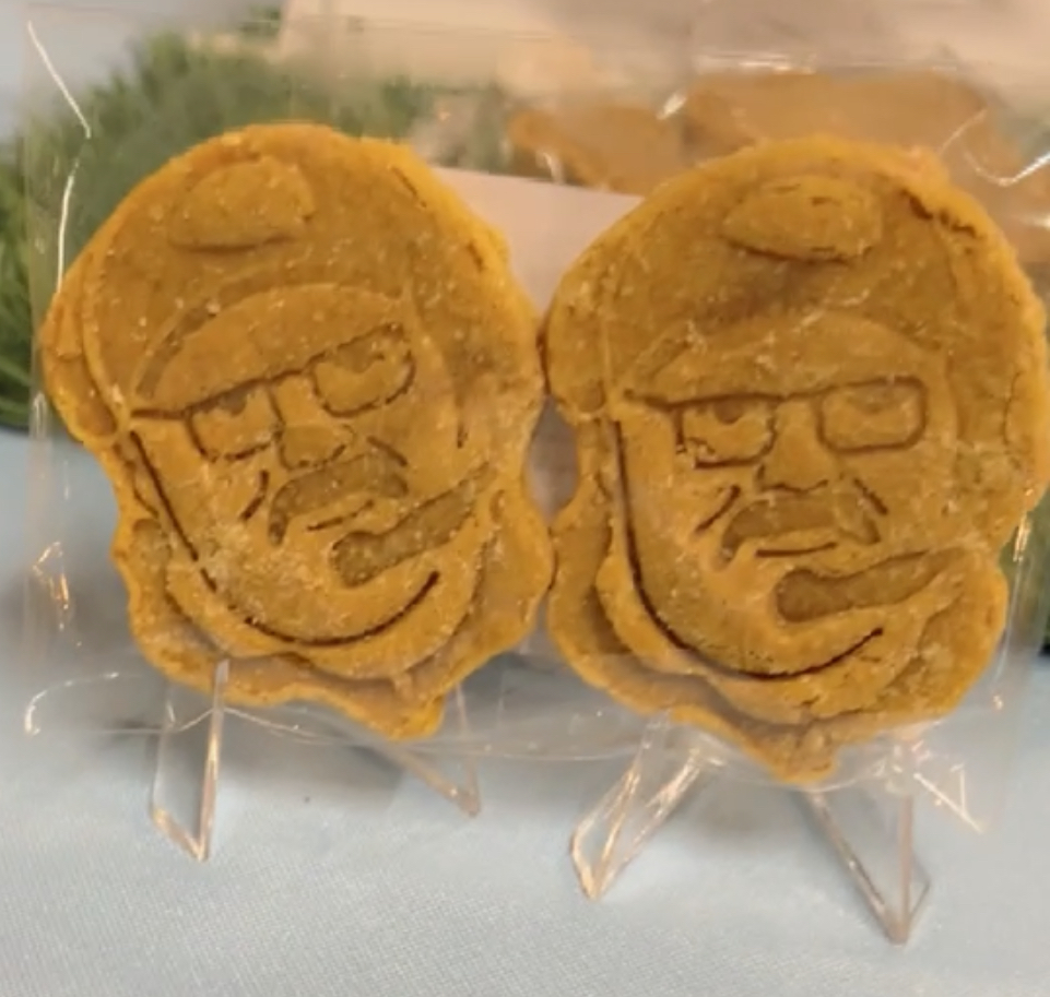 Two cookies with a face wearing glasses and a hat, wrapped in clear plastic, on a light surface.