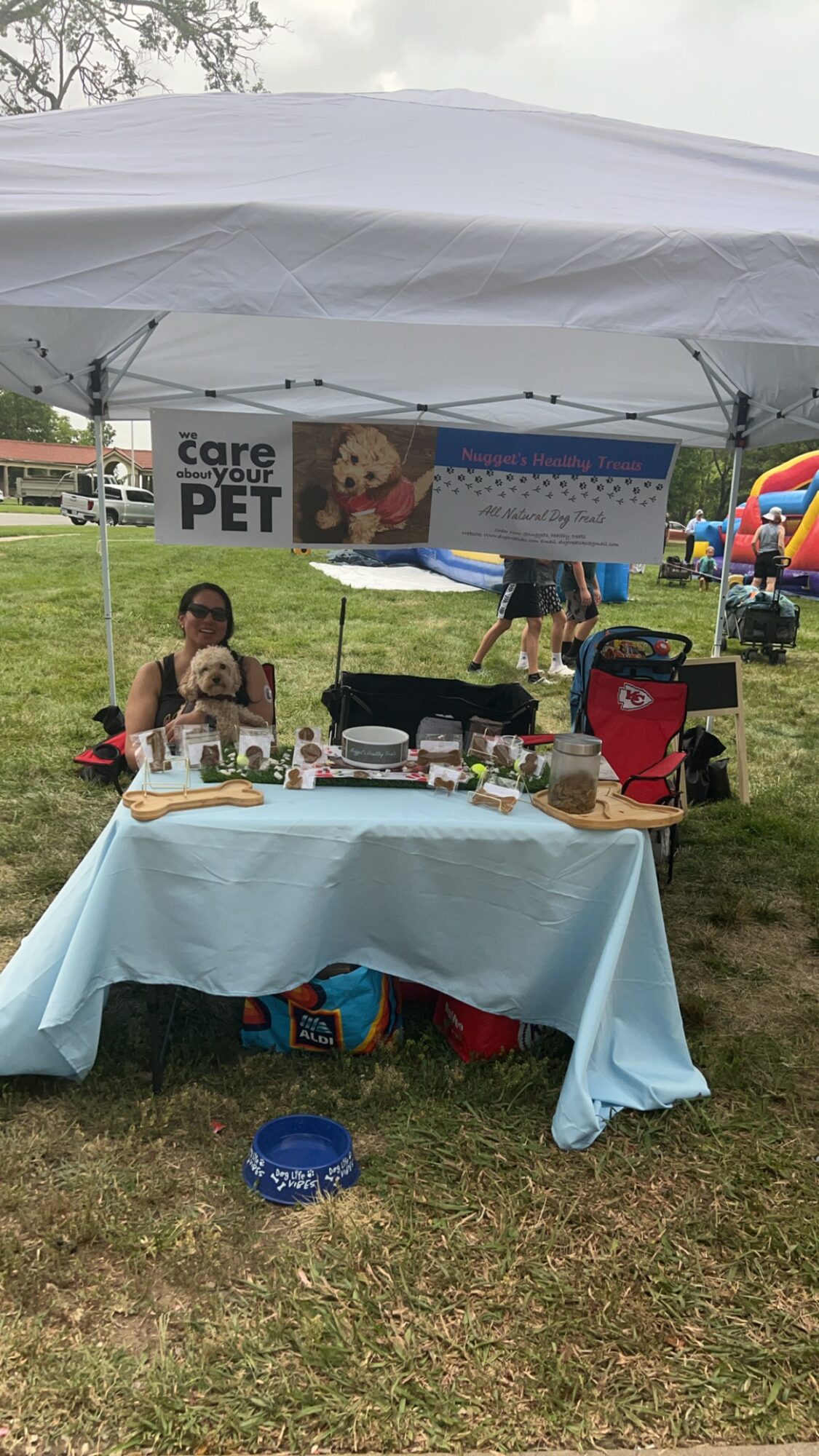 Table with pet care items under a canopy at outdoor event, with a banner and inflatable slide in background.