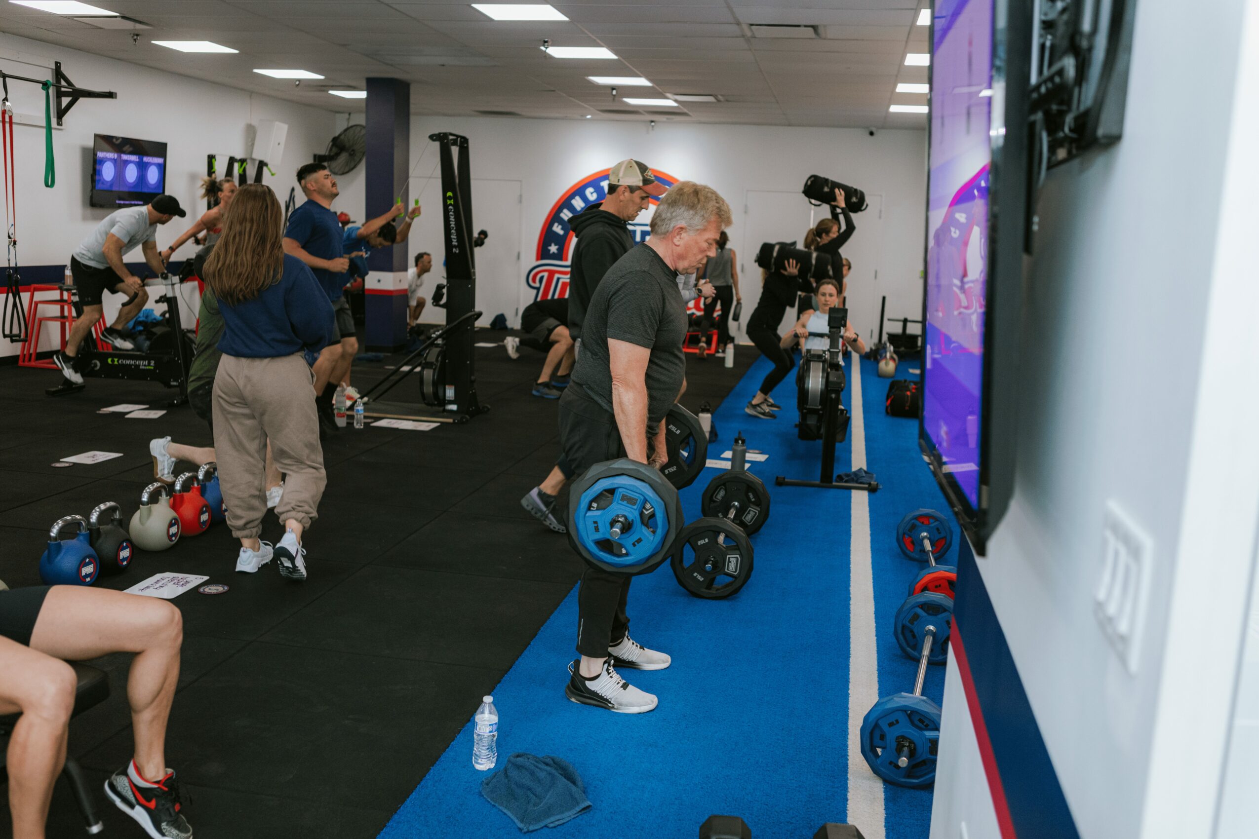 People exercising with weights and equipment in a gym, some lifting, others preparing or resting, with a large screen on the wall.