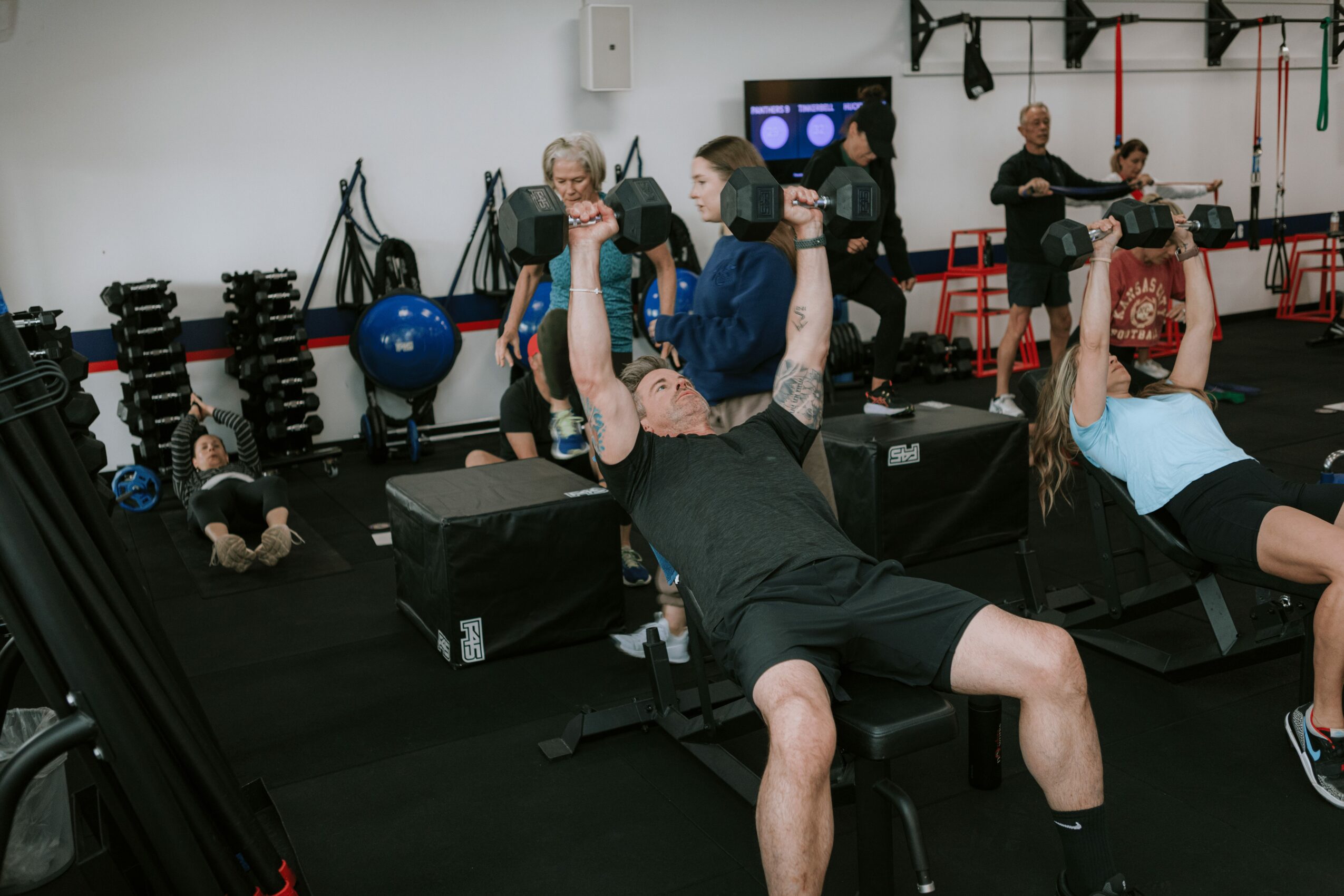 People exercising in a gym with weights, mats, and fitness equipment, some lying on benches and others standing or sitting.