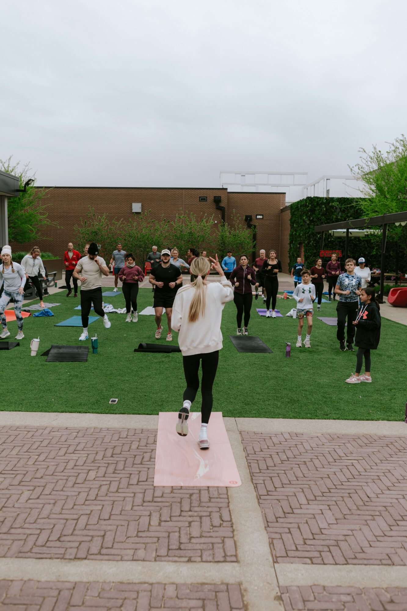 Person standing on a pink mat facing a group of people outdoors on a grassy area, with a brick building in the background.