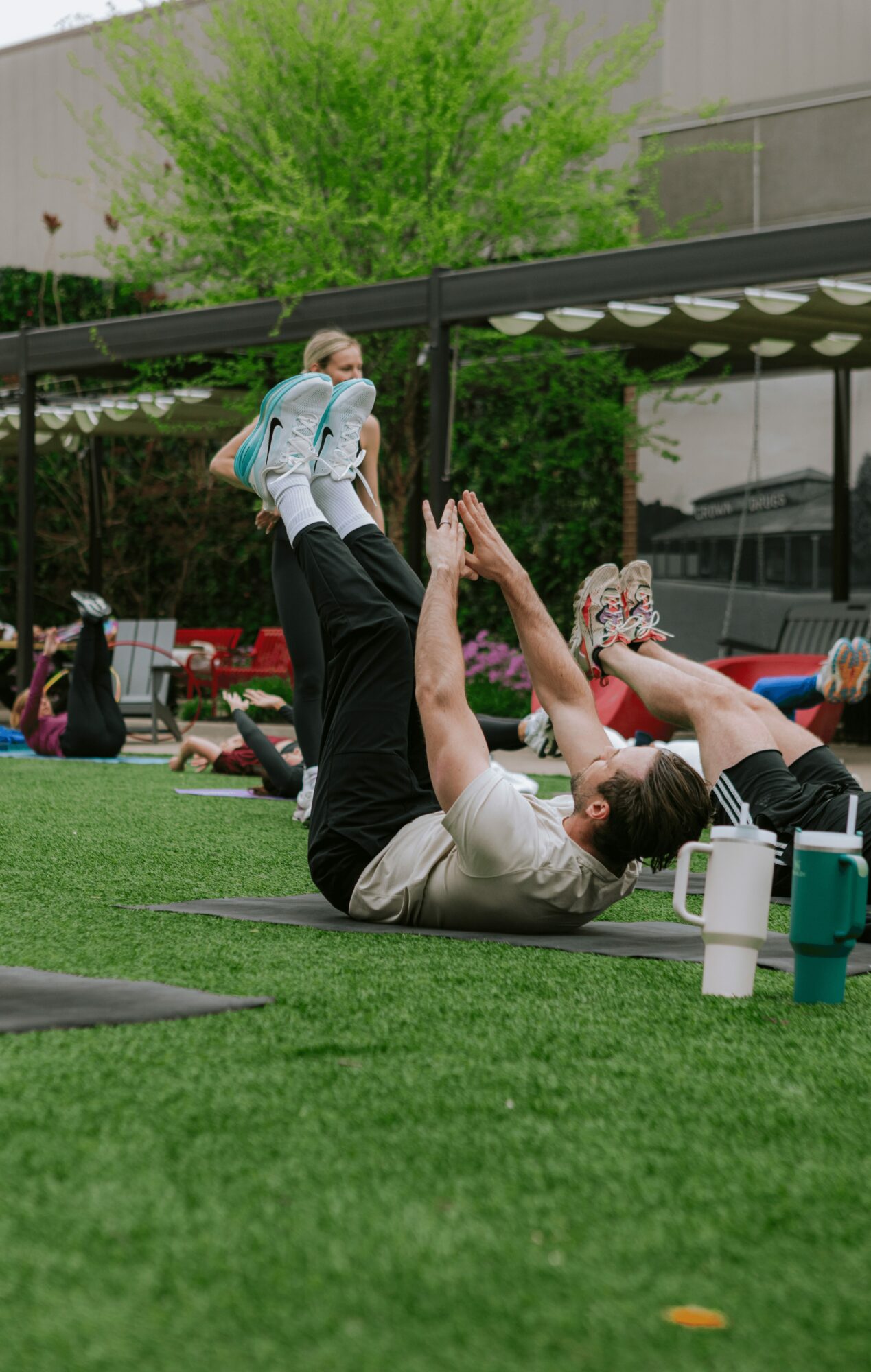 Person lying on grass with legs raised, holding a child in the air outdoors, surrounded by others and trees.