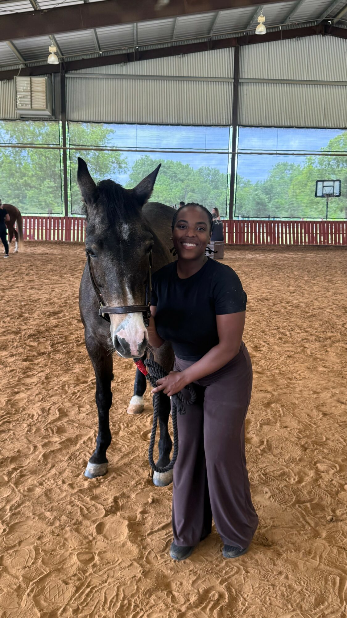 Person standing next to a horse inside an indoor riding arena, smiling, with a view of trees outside.