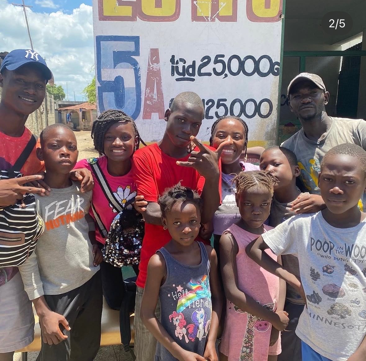 Group of children and adults outdoors, smiling, with a sign in the background displaying numbers and text.