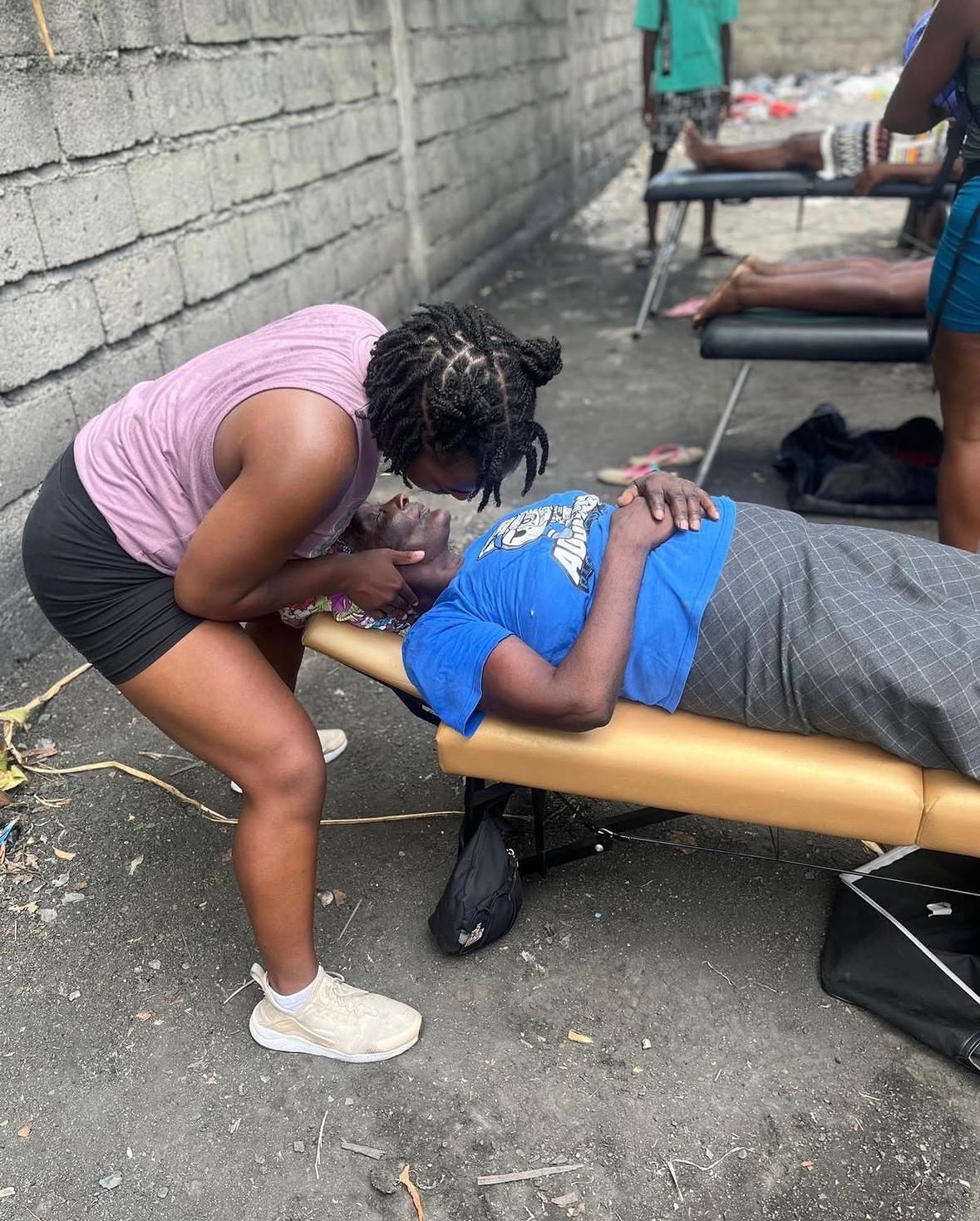 A woman in a pink top and black shorts leans over a person lying on a yellow medical bed outdoors, with a brick wall in the background.