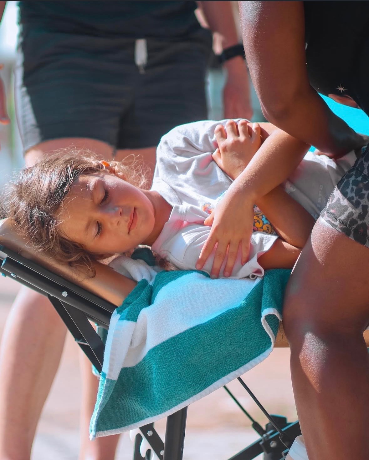 Young girl lying in a stroller with eyes closed, being attended to by a person, outdoors on a sunny day.