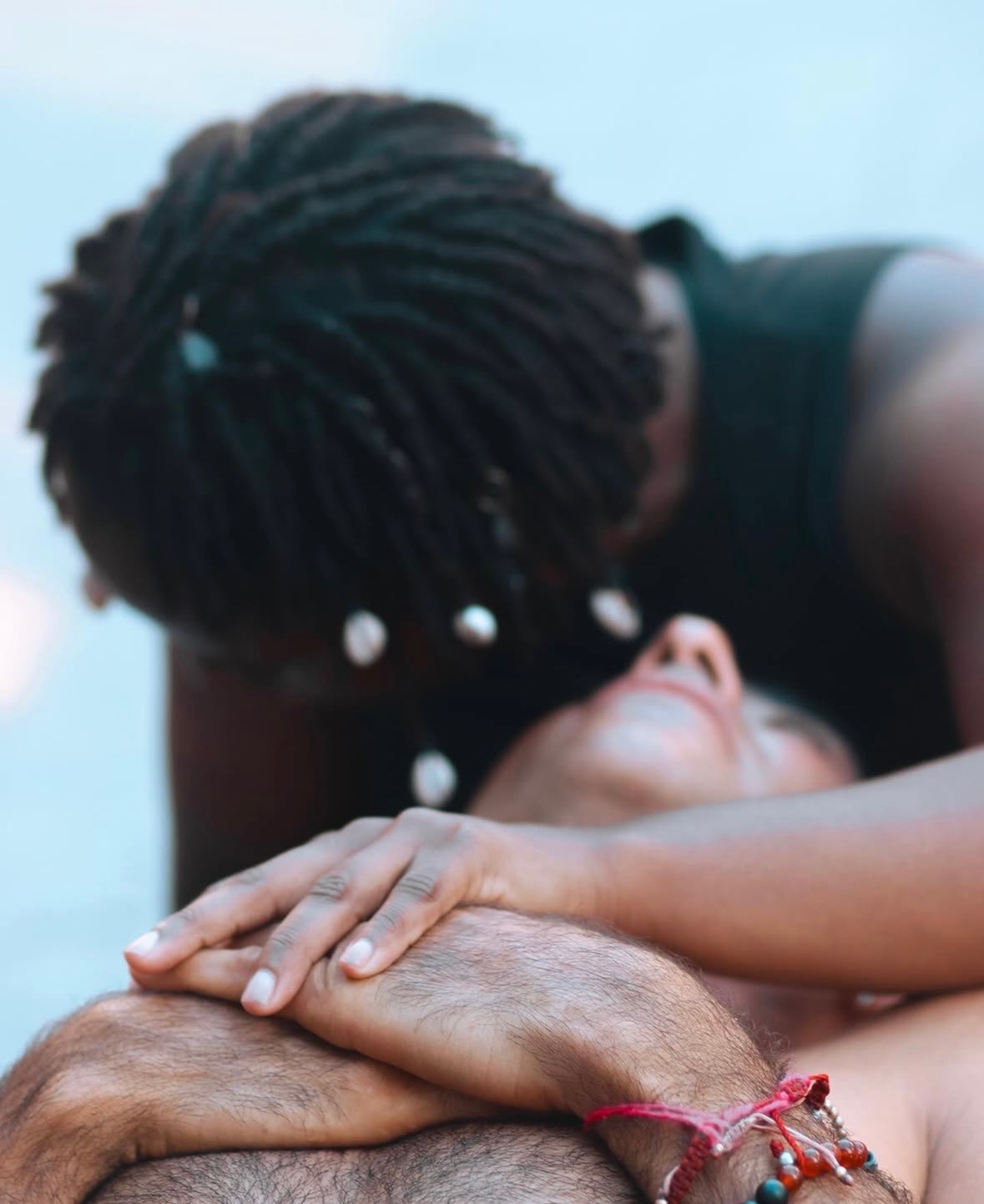 Two people lying close together, one with dreadlocks leaning over the other, who has their eyes closed and hands on their face.