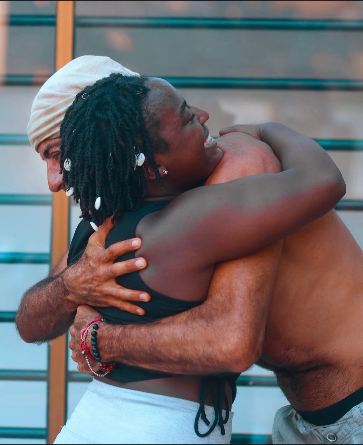 Two people hugging, one with dreadlocks and the other wearing a white cap, outdoors with a metal fence background.