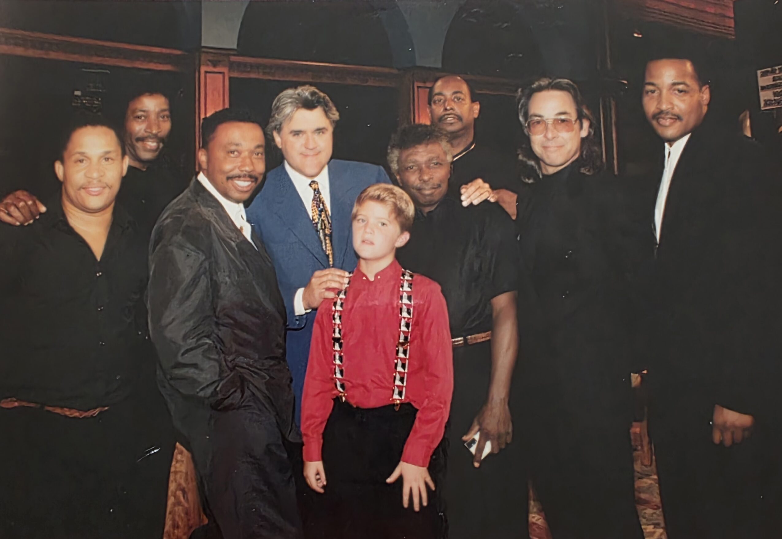 Group of nine people standing together indoors, smiling, with a dark background and red lighting accents.