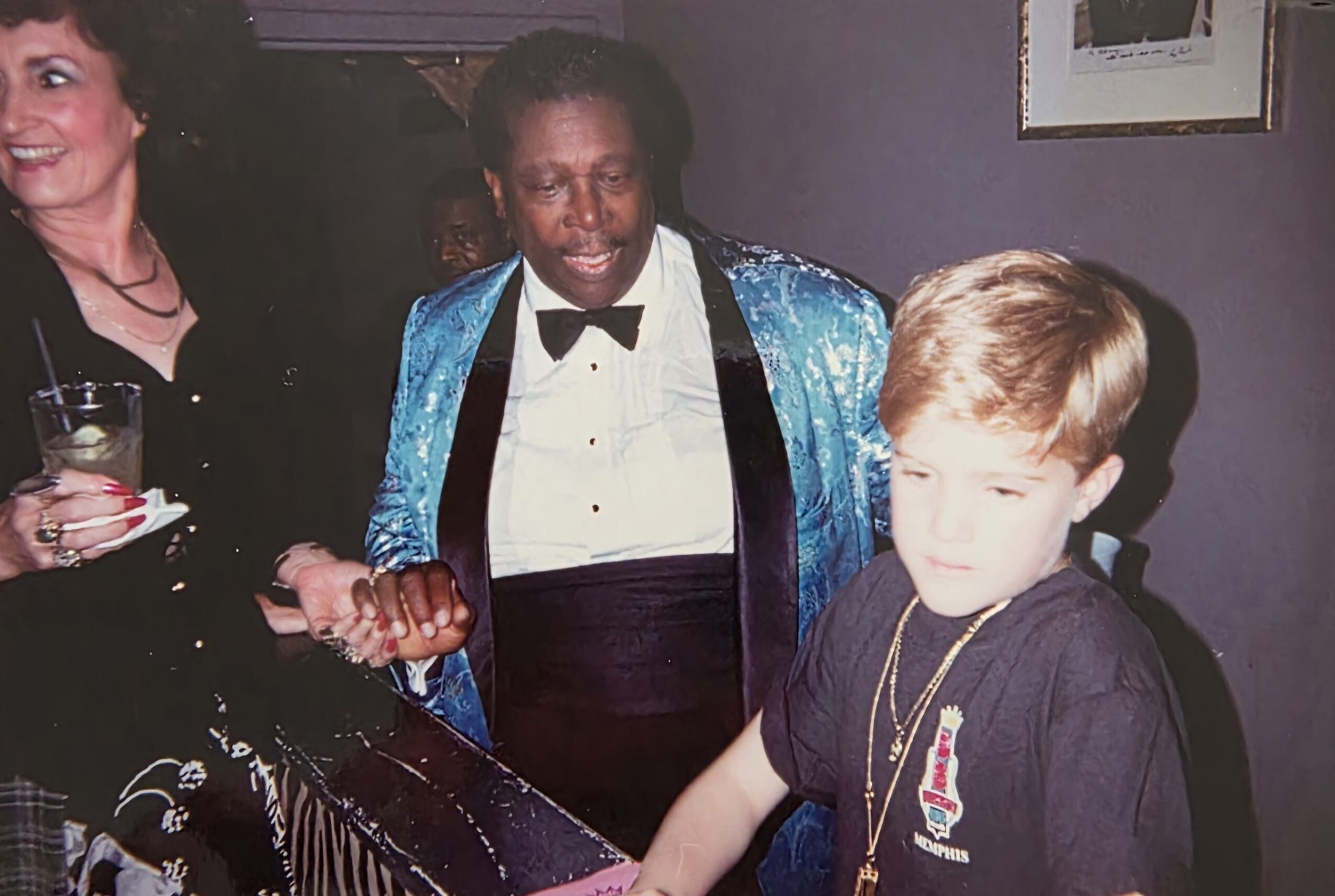 Person in a shiny blue jacket and bow tie smiling with a young boy at a bar or counter.