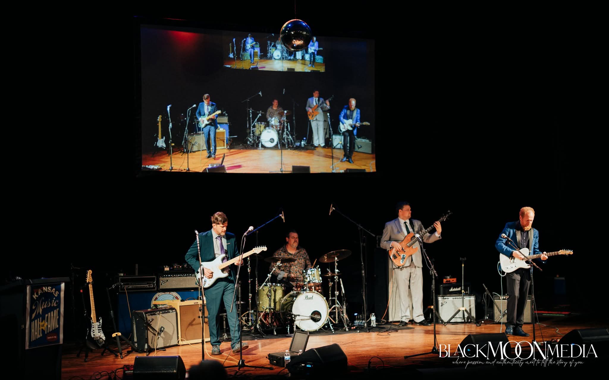 Band performs on stage with four musicians, drums, and a large screen above showing the performance, in a concert setting.