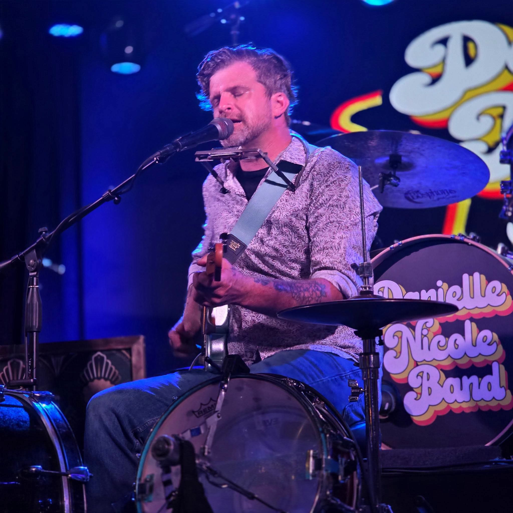 Musician playing guitar and singing into microphone on stage with drums and colorful background.