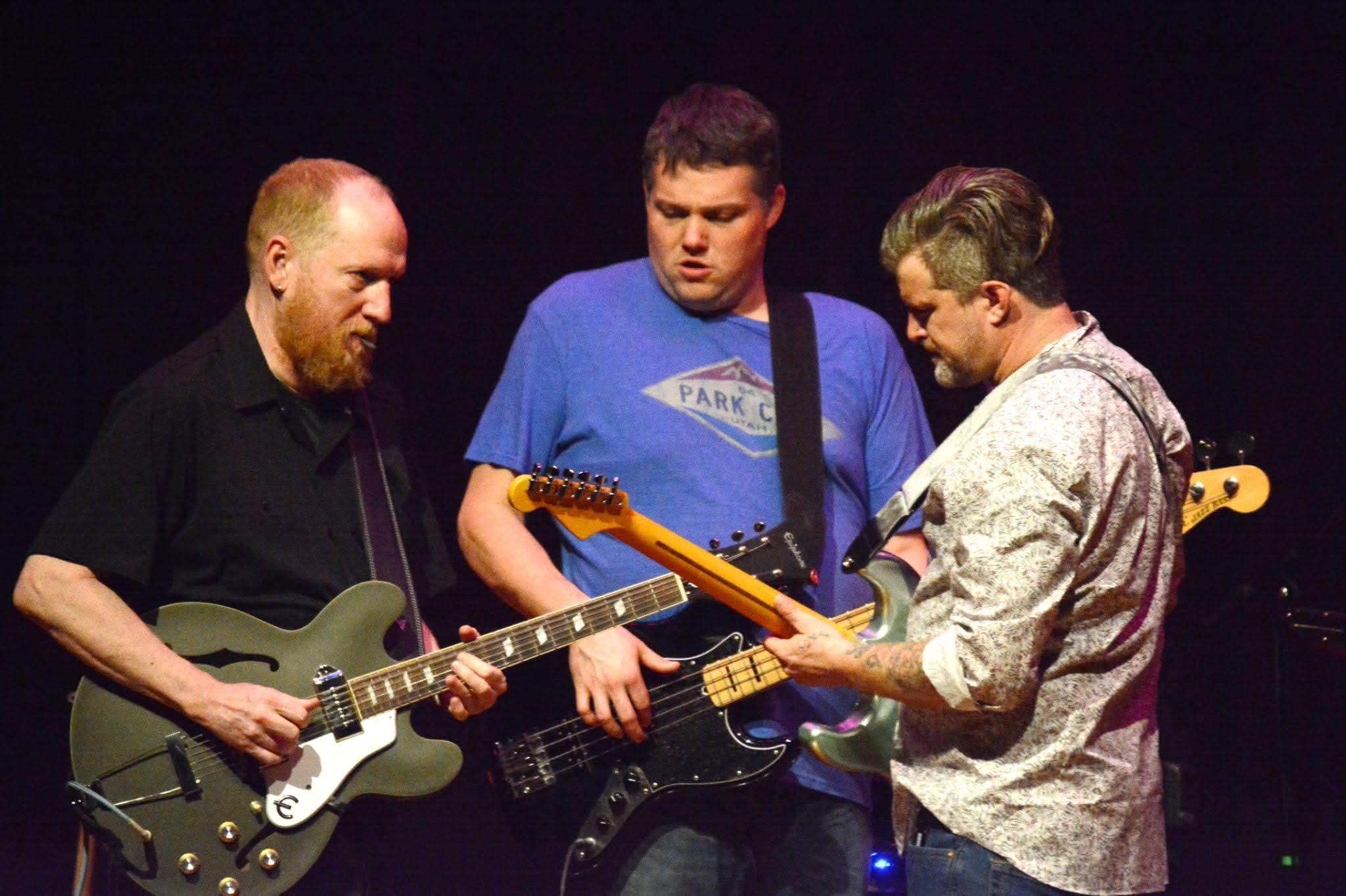 Three men playing electric guitars on stage, focused on their instruments, with a dark background.
