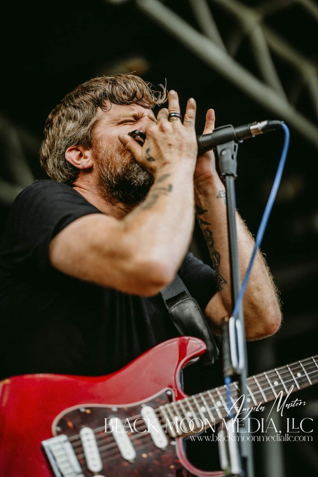 Male musician singing into microphone with guitar, eyes closed, in a dark stage setting.