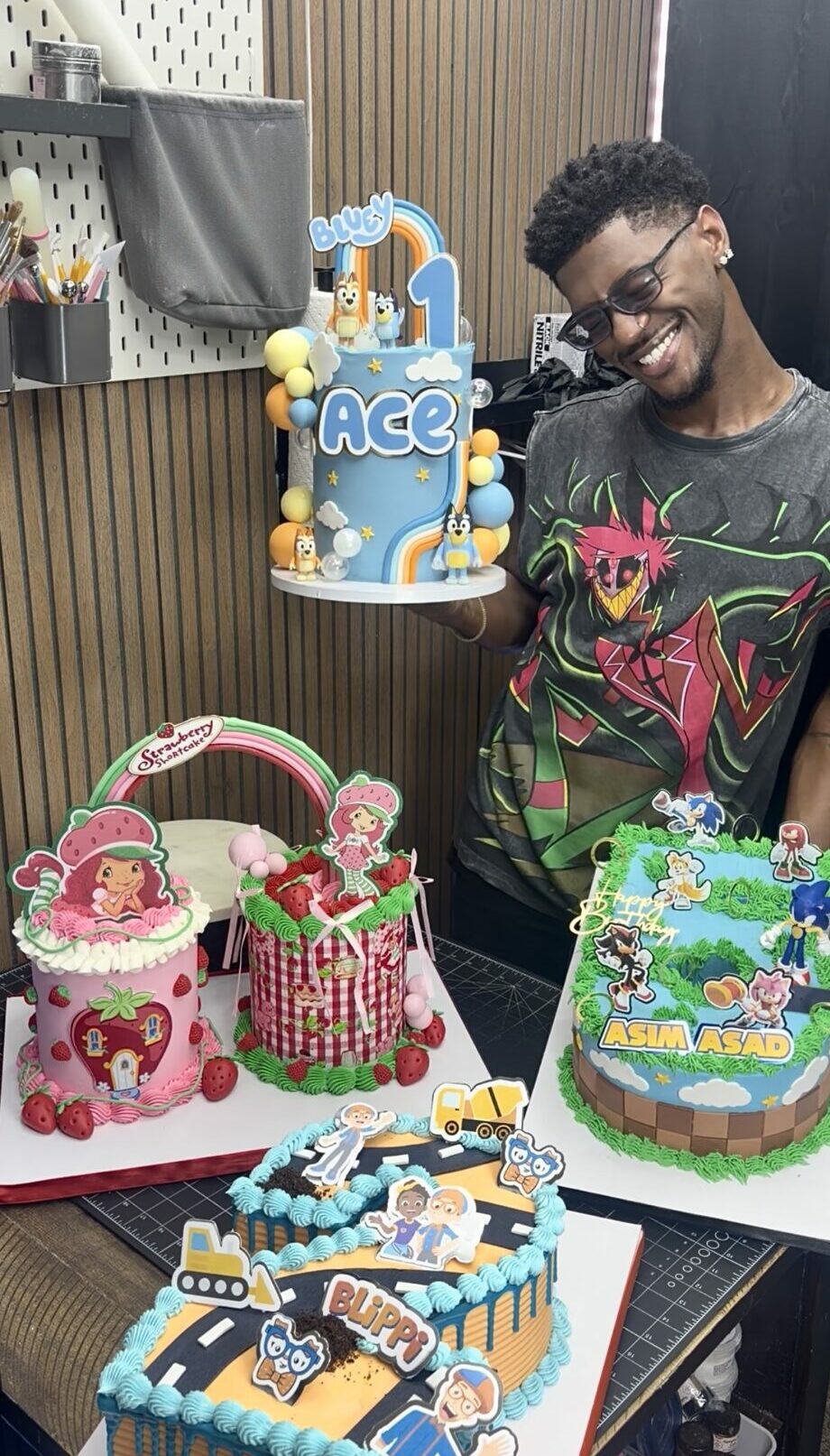 Young man with glasses smiling next to colorful decorated birthday cakes, including one with the name 'Ace' and a train theme.