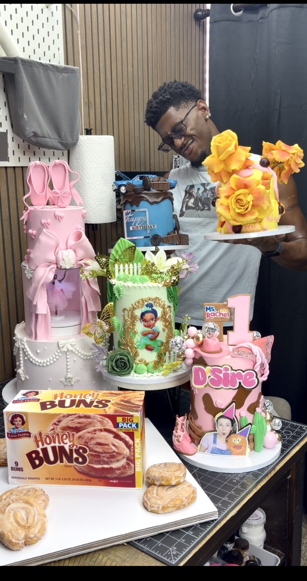 Young man with glasses holding a birthday cake with yellow flowers, surrounded by decorated cakes and snacks on a table.