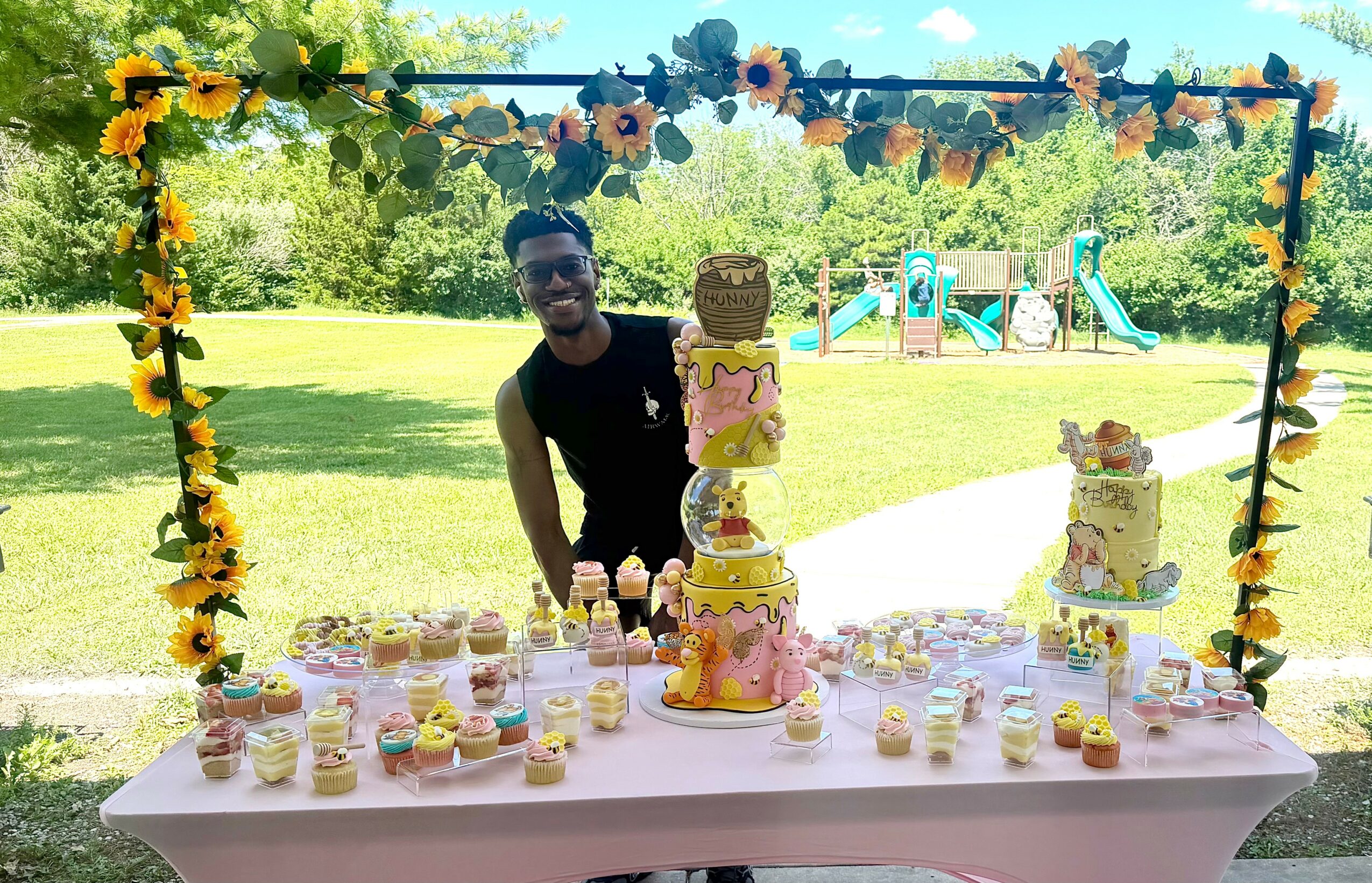 Person standing behind a table with cupcakes and a tiered cake outdoors, decorated with a sunflower and yellow theme.