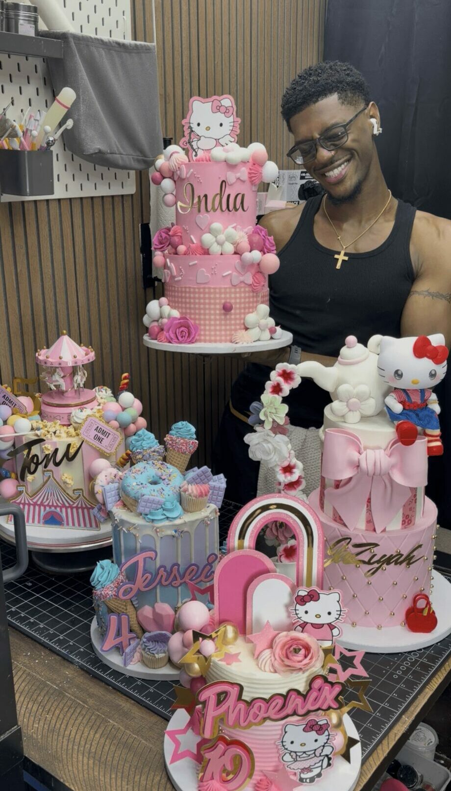 Smiling man with glasses and a black tank top behind a table of colorful decorated cakes and desserts.