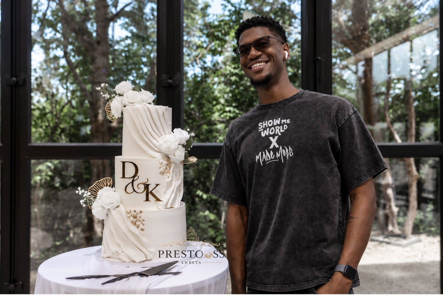 Smiling person standing next to a decorated cake with trees and glass windows in the background.